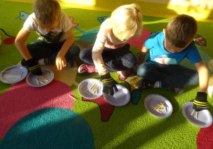 Trójka dzieci z założonymi rękawiczkami przekłada wykałaczki z jednego talerzyka na drugi